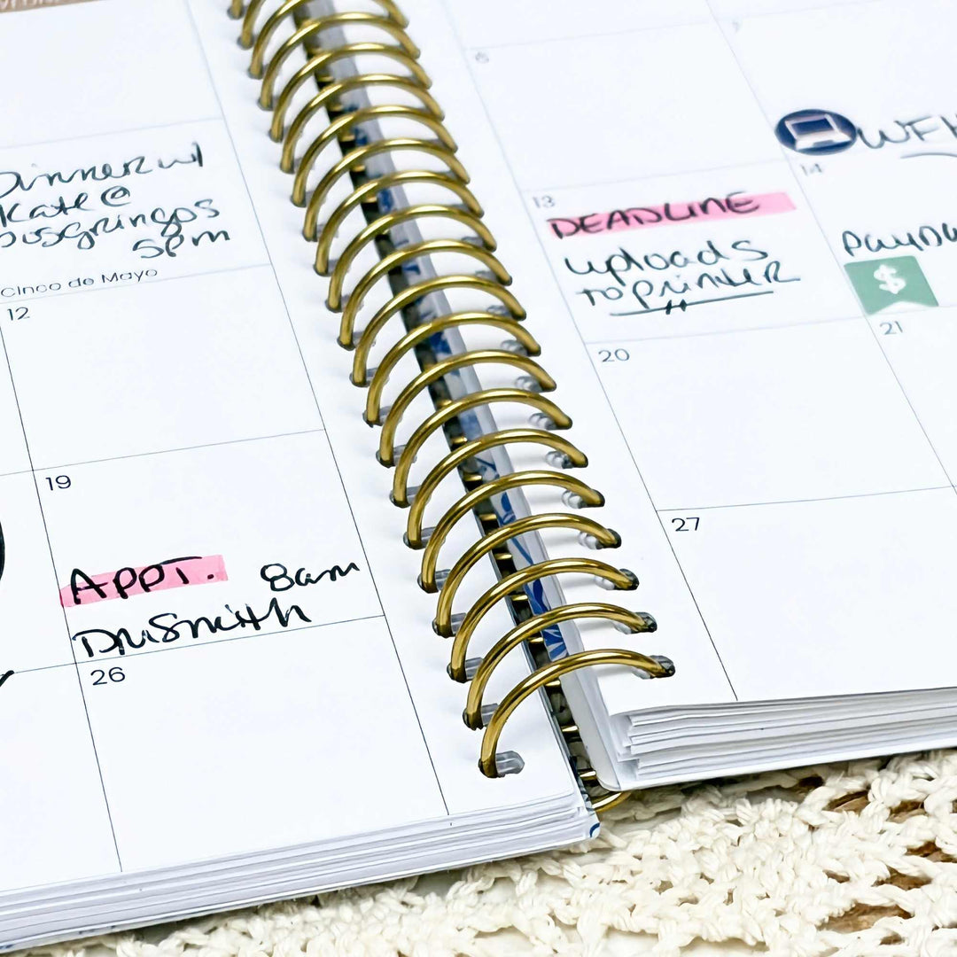 A close-up of a spiral-bound planner or calendar showing handwritten notes on the pages, with a gold-tone spiral binding and white paper pages.