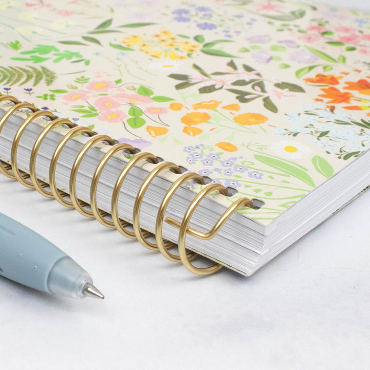 A close-up of a spiral-bound notebook with a gold wire coil and a pastel floral cover pattern, shown on a white surface with a light blue pen partially visible in the foreground.