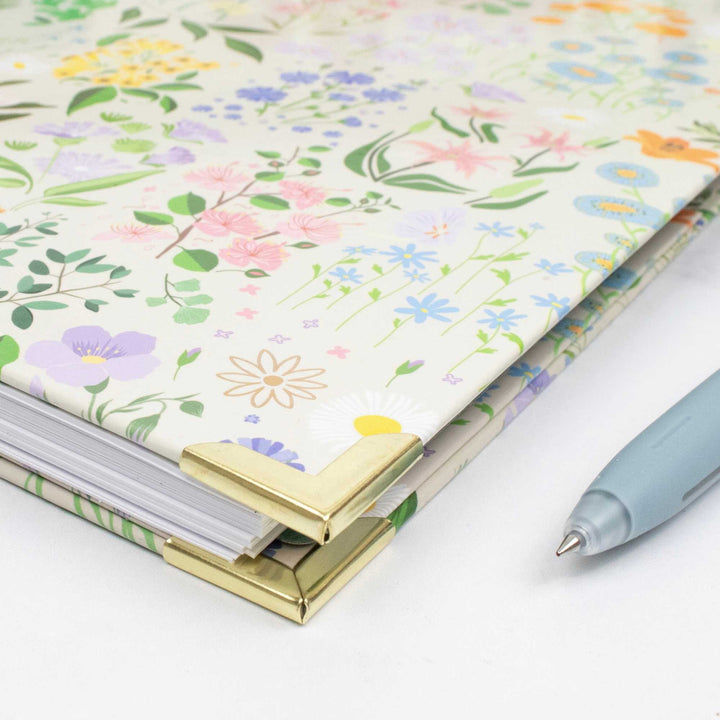 A floral-patterned notebook with gold metal corner protectors and a light blue pen on a clean white surface.