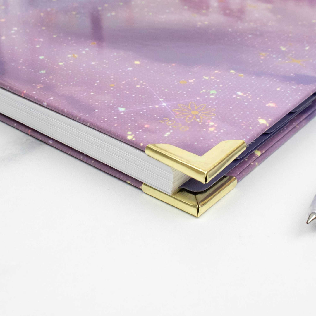 A close-up of a hardcover notebook or journal with a lavender-purple glittery cover, white pages, and shiny gold corner protectors on a light surface.