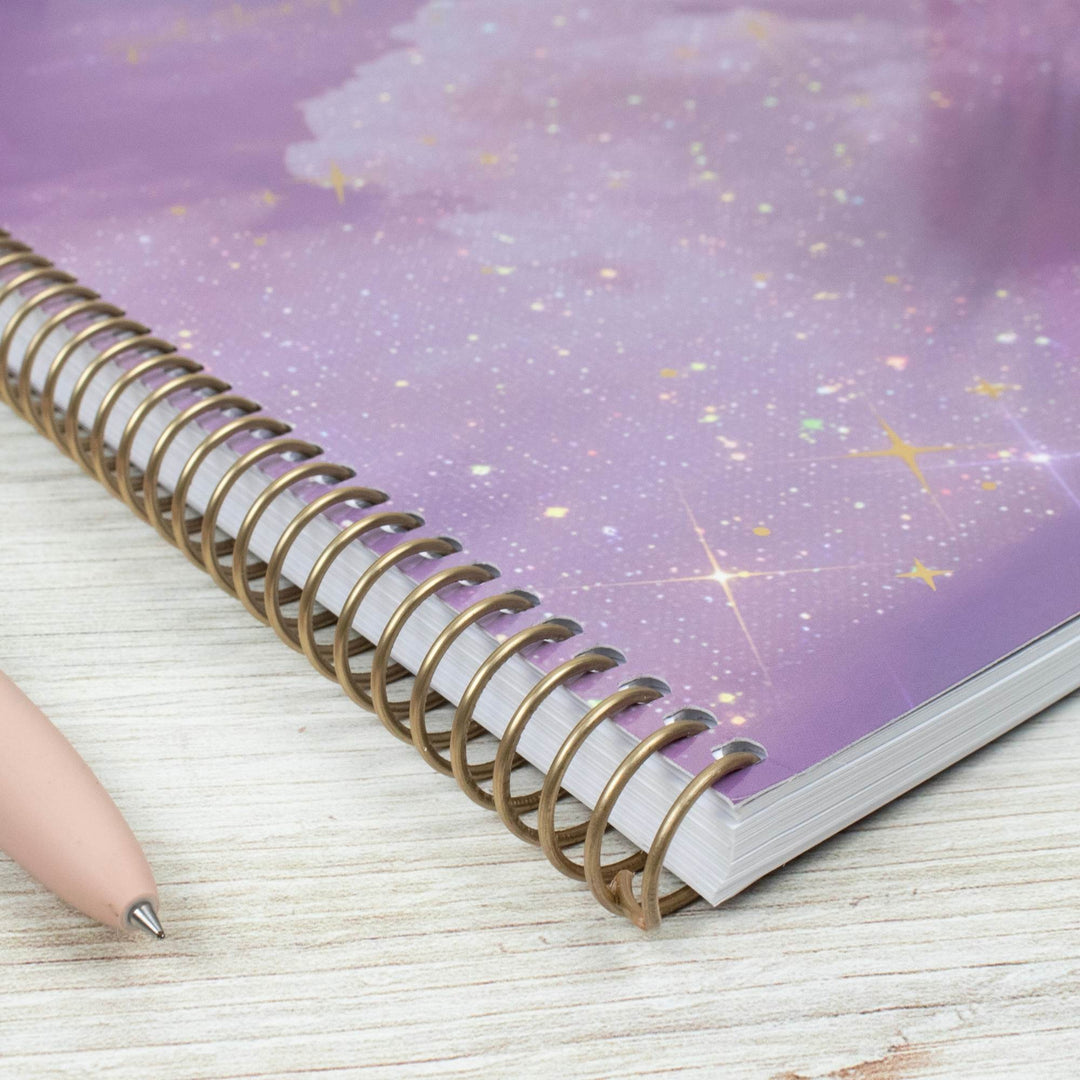 A spiral-bound notebook with a purple galaxy cover, resting on a light wood surface next to a pink pen.