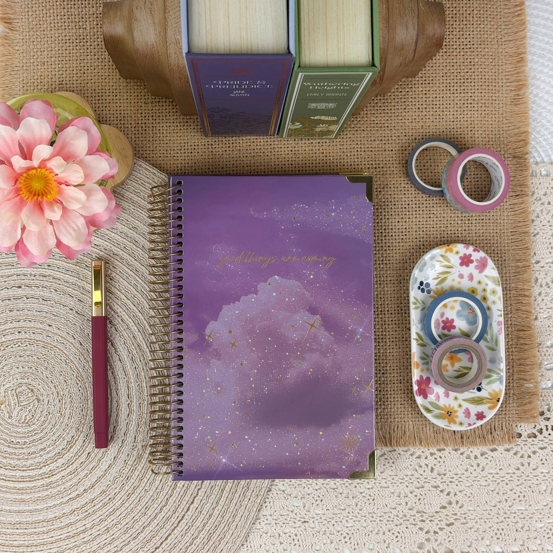 A purple spiral-bound notebook with a galaxy print and gold glitter accents, styled in a flat-lay arrangement with a pink flower, a pen, books, washi tape, and a small floral container on a burlap and lace background.