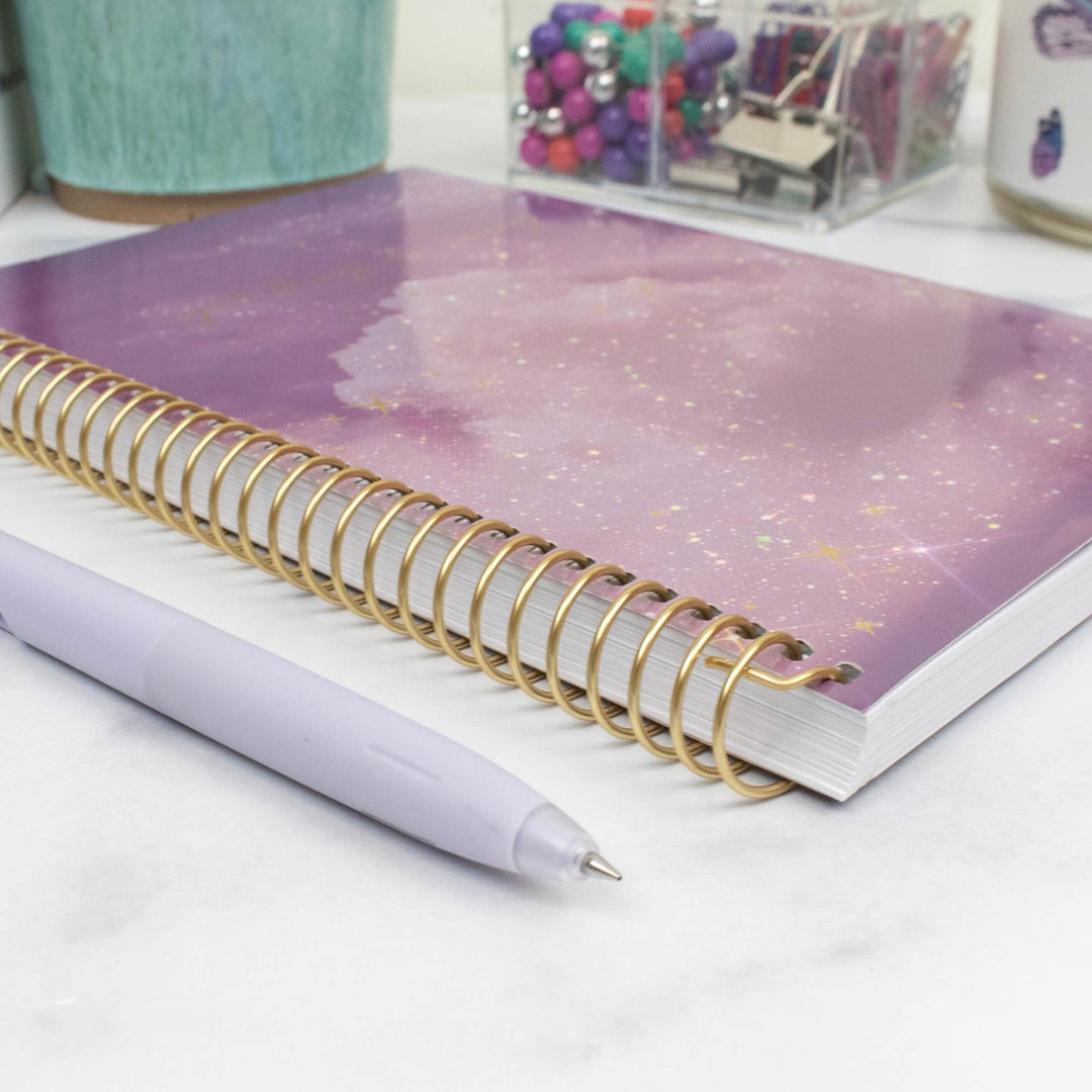 A spiral-bound notebook with a purple galaxy-patterned cover and gold wire binding, shown on a white marble-like surface with a lavender pen in front and small desk accessories blurred in the background.