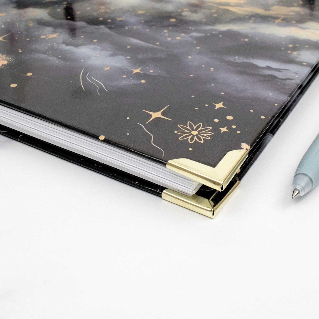 A close-up of a hardcover notebook or journal with a black celestial cover design featuring gold stars, floral motifs, and gold corner protectors, shown on a white surface next to a light blue pen.