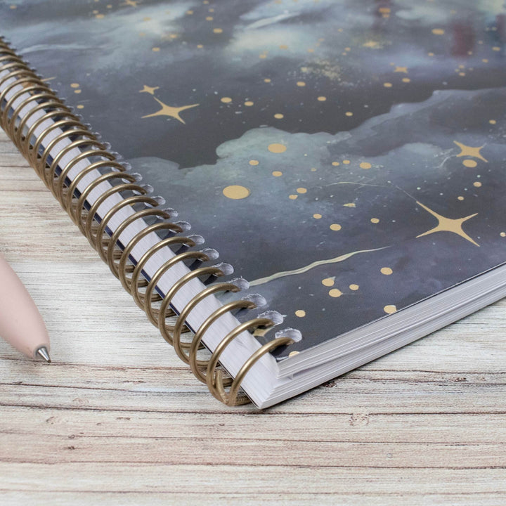 A spiral-bound notebook with a dark blue and gray galaxy cover featuring gold stars and clouds, shown on a light wood surface next to a pink pen.