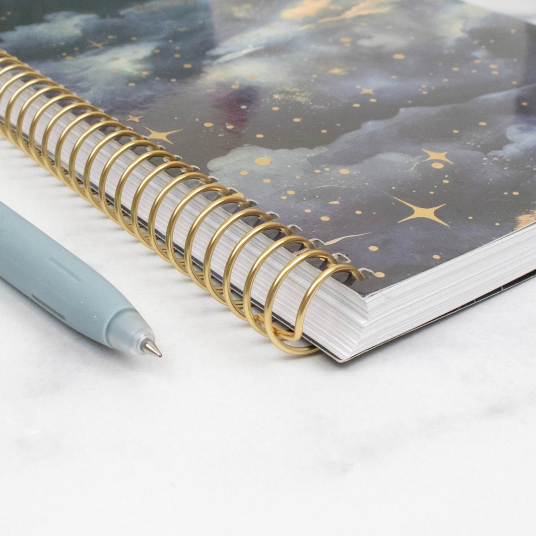 A spiral-bound notebook with a dark galaxy-patterned cover and gold wire binding, shown on a white marble-like surface next to a light blue pen.