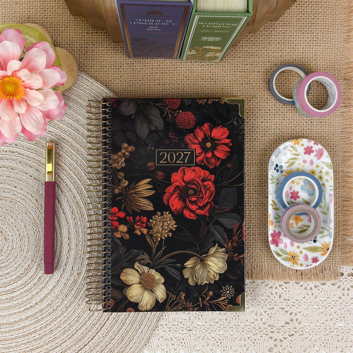 A 2027 spiral-bound floral planner lies on a burlap mat, surrounded by stationery props including a pink pen, washi tapes, a small dish with colorful tape, and flowers.