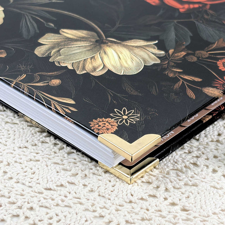 A close-up of a hardcover notebook or journal with a black cover decorated with large gold and orange floral illustrations, resting on a cream-colored textured knit blanket. The notebook has polished gold metal corner protectors.