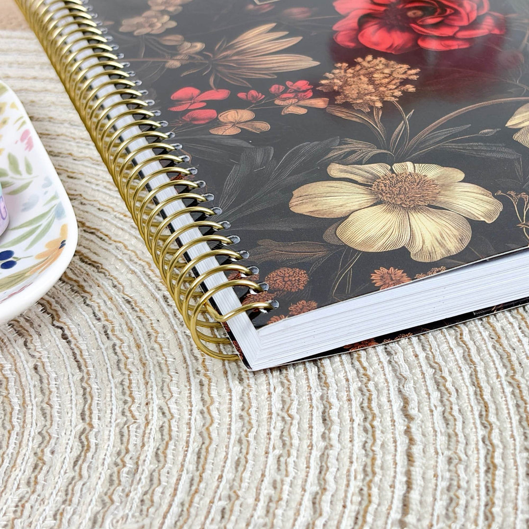 A spiral-bound notebook with a dark floral cover lies on a textured beige woven placemat. The cover features large cream flowers, red blossoms, and botanical illustrations.