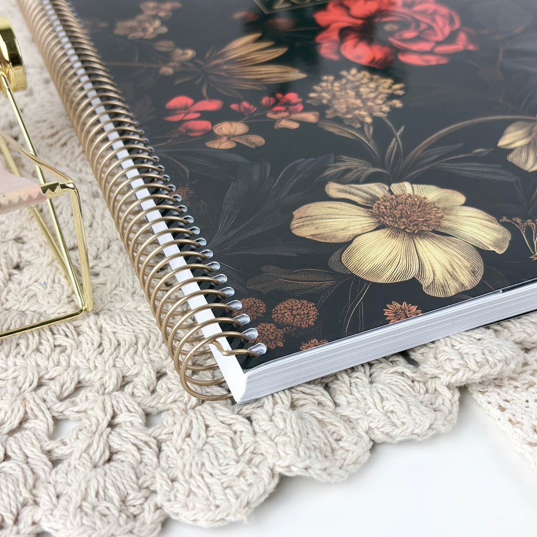 A spiral-bound notebook with a dark floral cover, styled on a cream crochet blanket next to a gold geometric stand.