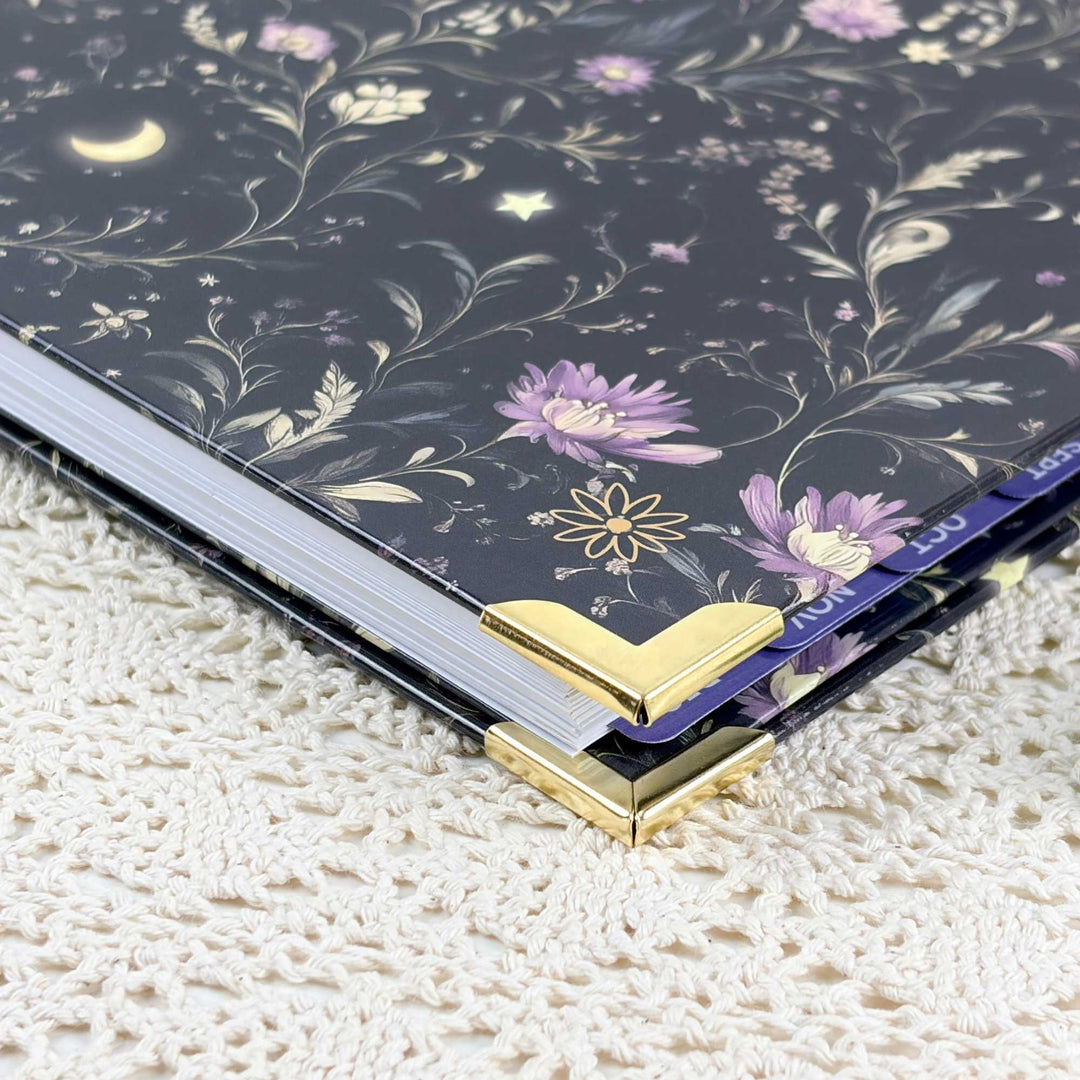 A close-up of a decorative notebook or journal with a dark navy cover featuring a celestial floral pattern of purple flowers, gold stars and moons, and gold corner protectors, resting on a cream-colored textured knit blanket.