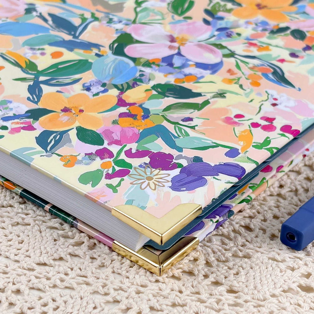 A close-up of a floral-patterned notebook or journal with gold metal corner protectors, resting on a cream-colored textured knit blanket. The cover features a colorful watercolor-style floral print with pink, purple, blue, green, and orange flowers on a pale yellow background.