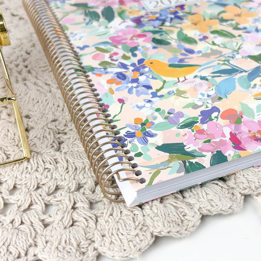A spiral-bound notebook with a colorful floral and bird pattern on the cover, placed on a beige crocheted mat next to a gold metal stand.