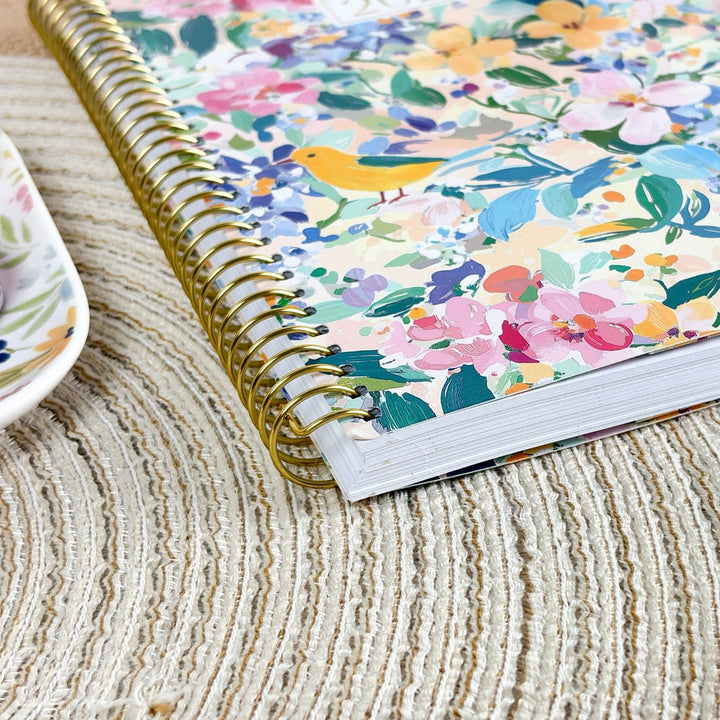 A spiral-bound notebook with a colorful floral and bird patterned cover, resting on a textured beige woven placemat next to a matching floral teacup.
