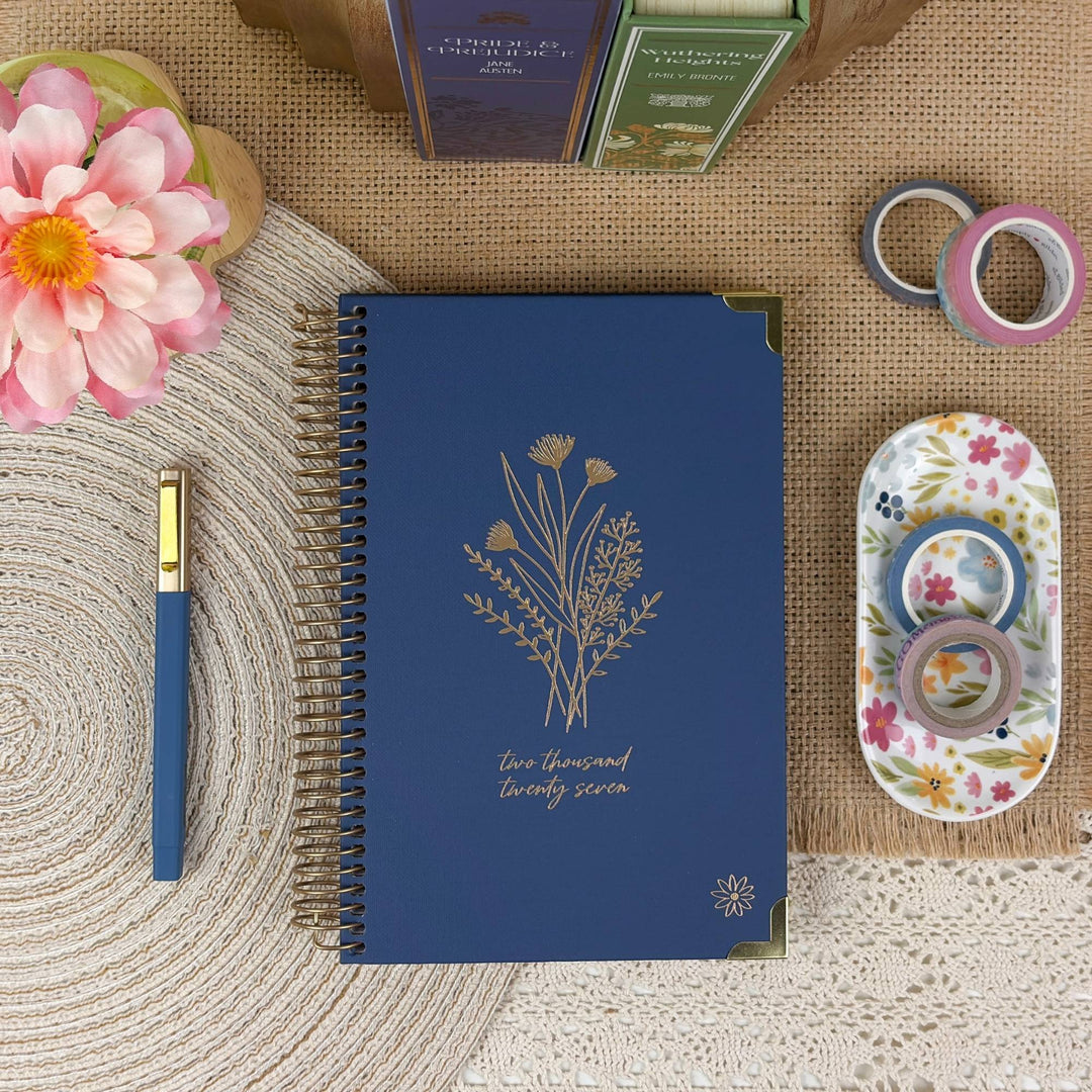 A navy blue spiral-bound planner or journal with gold floral line art and the text 'two thousand twenty seven' lies on a woven beige mat. A matching blue pen with a gold top is placed beside it, along with decorative washi tape rolls in a floral holder and a pink flower in the corner.