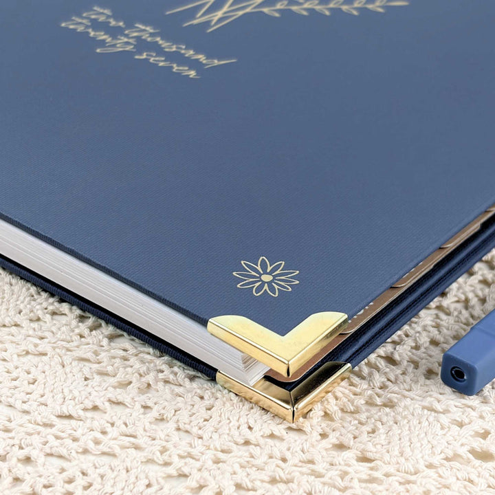 A close-up of a navy blue hardcover notebook or journal with gold foil lettering and a small gold flower emblem, resting on a cream textured knit surface. Gold corner protectors are visible on the edge.