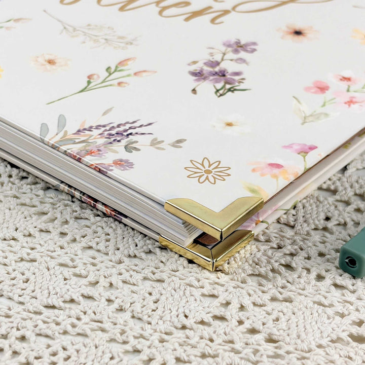 A close-up of a floral planner or notebook with gold corner protectors, resting on a cream-colored crocheted surface.
