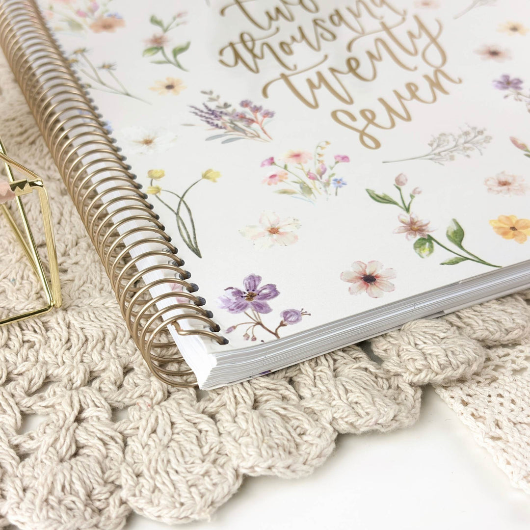 A spiral-bound planner or notebook with a white cover decorated with pastel floral illustrations and gold script text, styled on a cream crocheted mat with a small gold geometric prop.