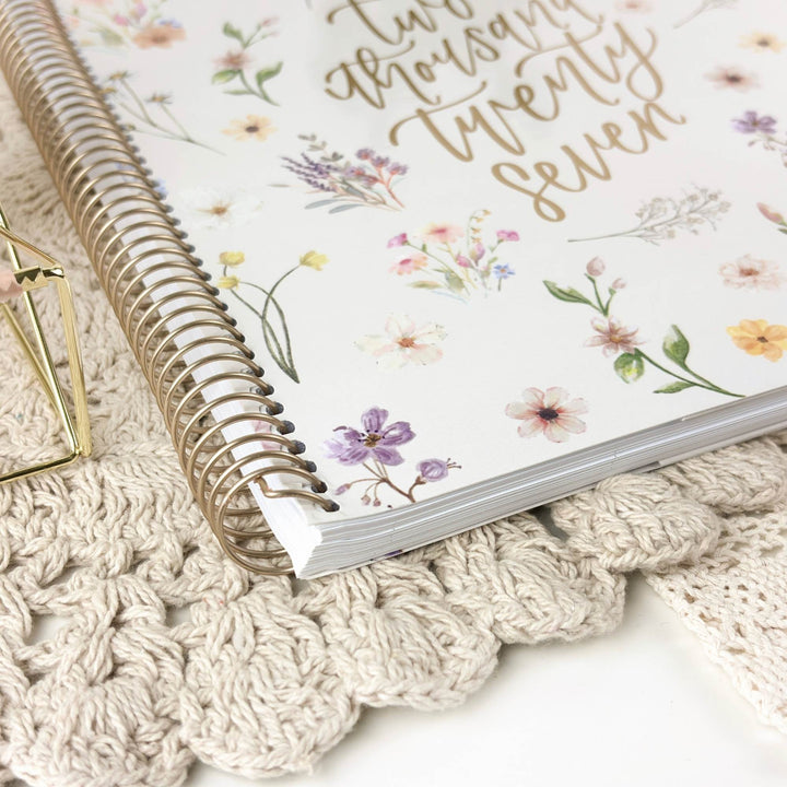 A spiral-bound planner or notebook with a white cover decorated with pastel floral illustrations and gold script text, styled on a cream crocheted mat with a small gold geometric prop.