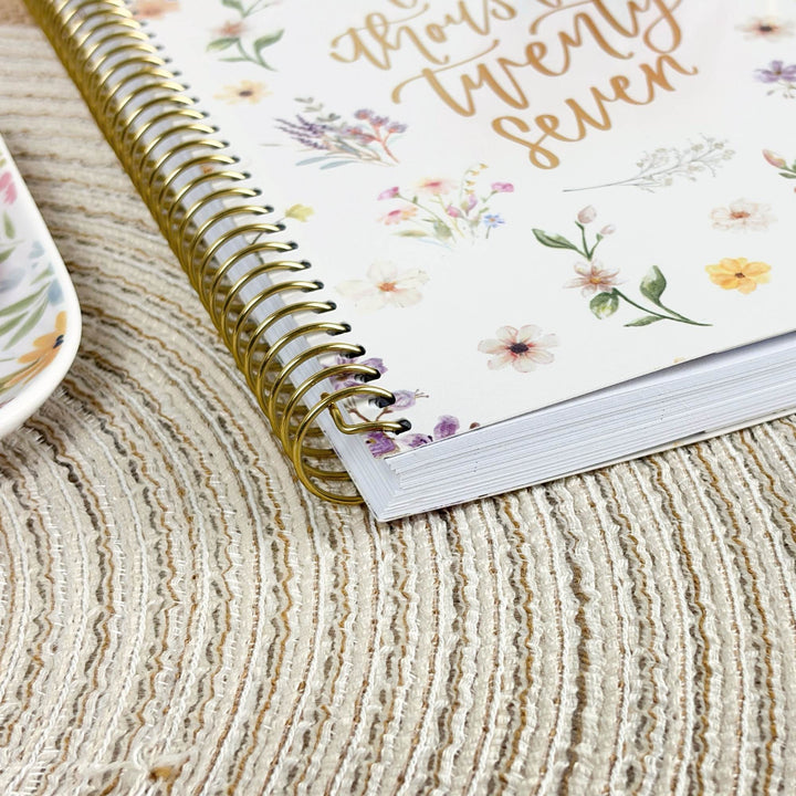 A spiral-bound planner or notebook with a white cover, gold wire binding, and delicate floral illustrations, placed on a textured beige woven surface.