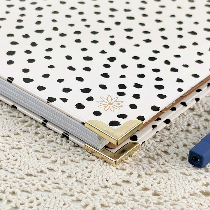 A hardcover notebook with a white cover decorated with black dalmatian-style spots and a small gold flower motif, resting on a cream-colored crocheted blanket. The notebook has gold metal corner protectors and is shown with a blue pen partially visible in the corner.