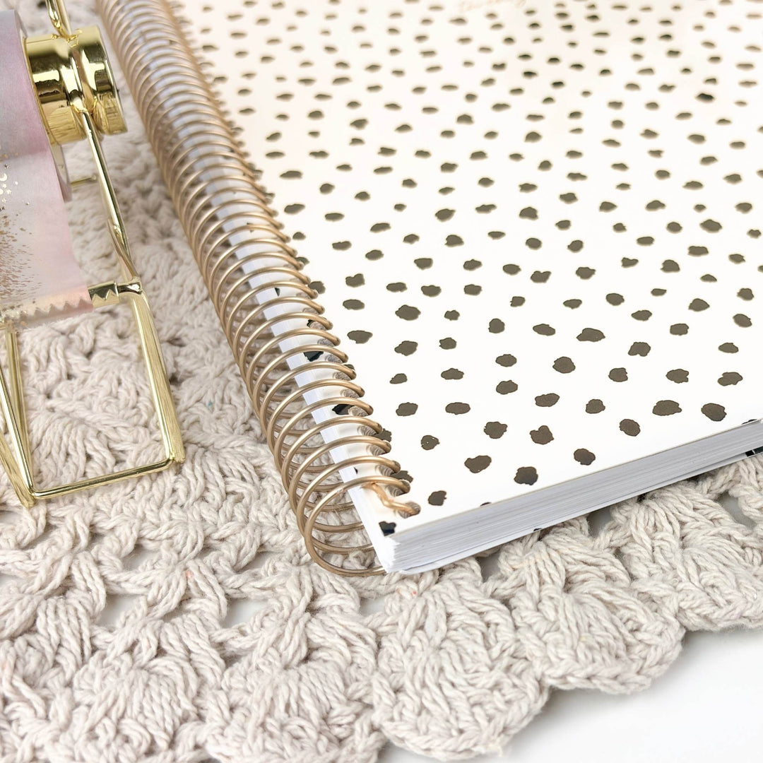 A spiral-bound planner or notebook with a white cover and small gold polka dots, photographed close up on a cream crocheted surface next to a gold-toned decorative object.