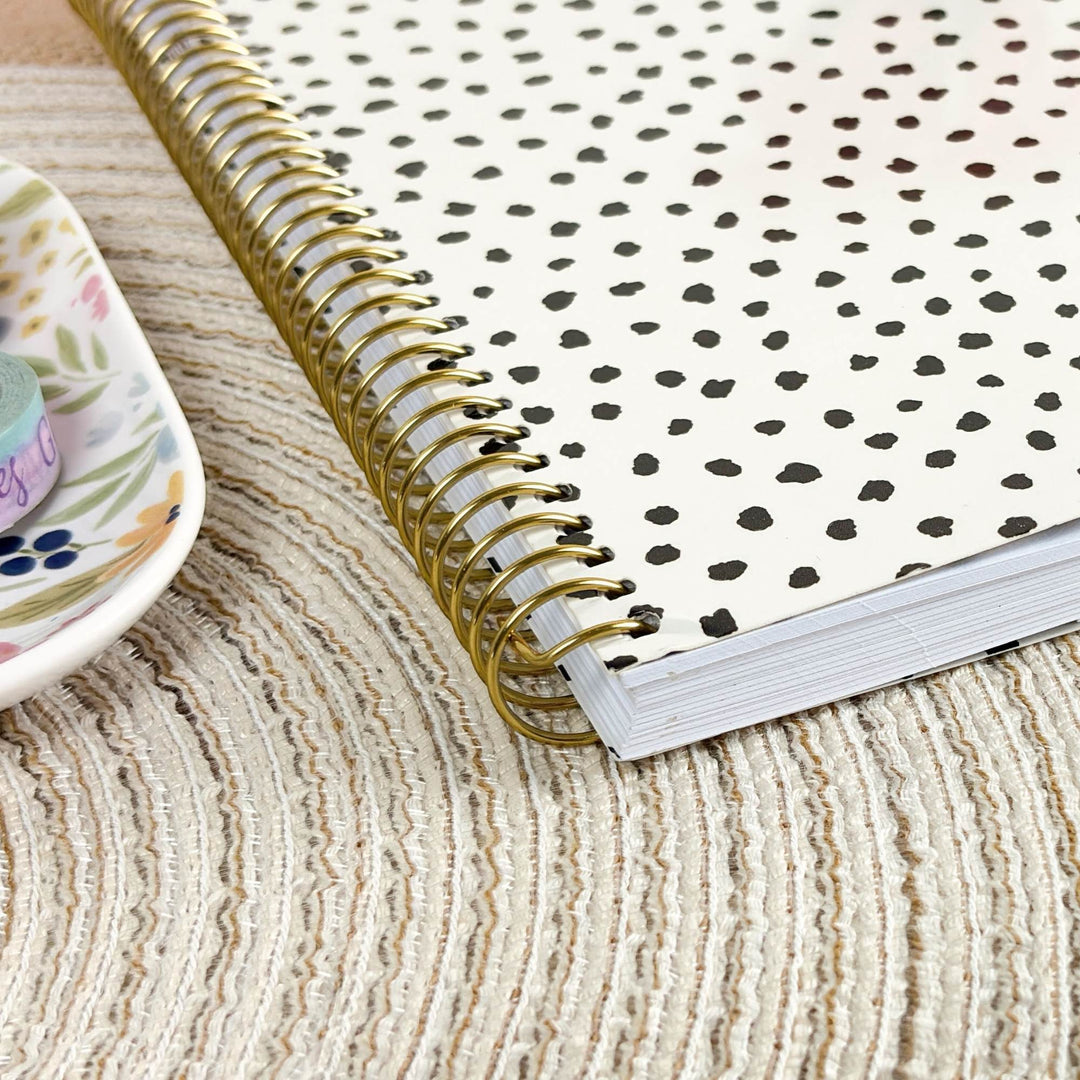 A close-up of a spiral-bound notebook with a gold wire binding and an off-white cover covered in small black heart-like spots, placed on a woven beige rug next to a floral dish.