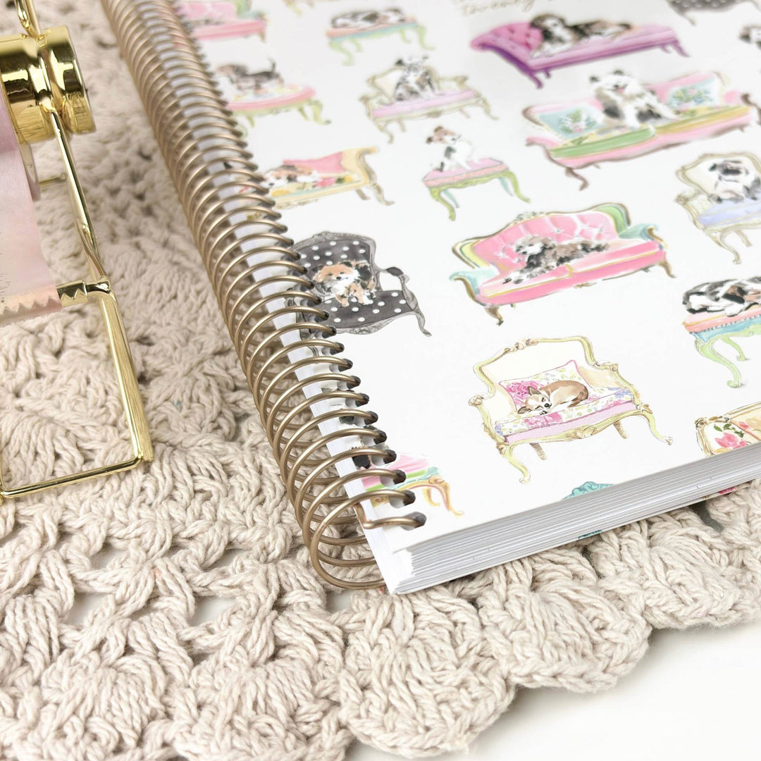 A spiral-bound notebook with a white cover decorated with colorful illustrations of dogs lounging in pink and green sofas, styled on a beige crochet blanket next to a gold metal object.