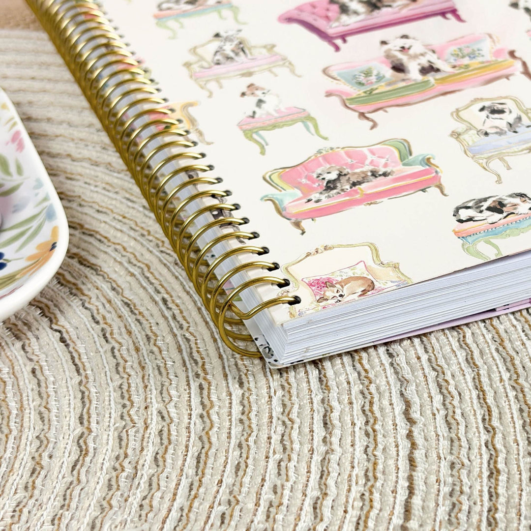 A spiral-bound notebook with a gold wire binding and a printed cover featuring illustrated cats lounging in pink and green sofas, photographed on a beige textured rug with a partial floral plate visible at the left edge.