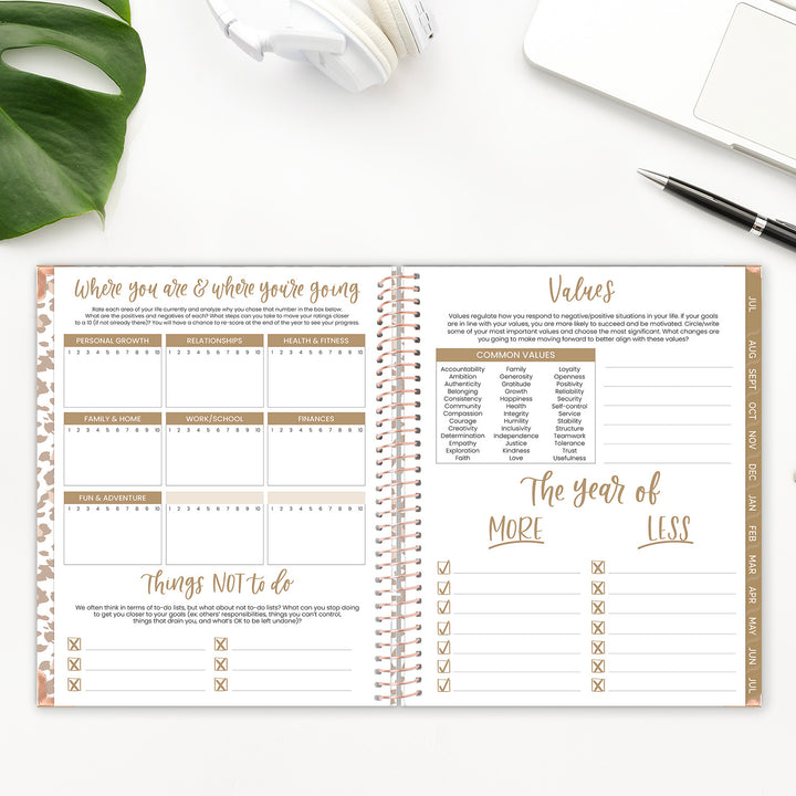 Spiral-bound planner with open pages on a white surface with a plant and pen.