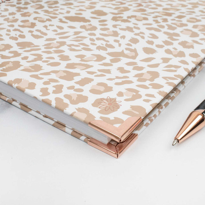A hardcover notebook with a beige and white leopard print cover and rose gold metal corner protectors, shown on a white surface next to a rose gold pen.