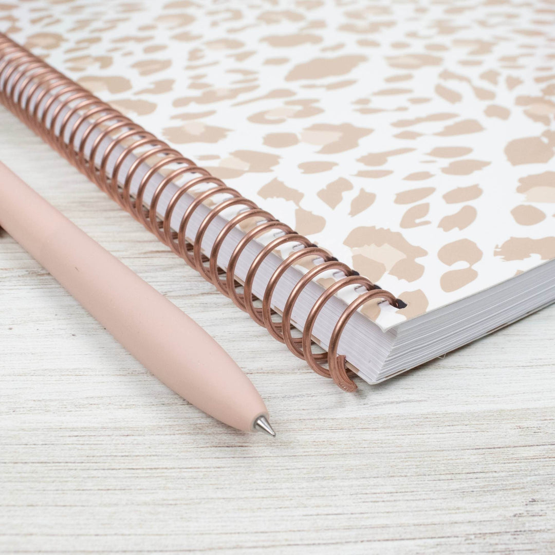 A spiral-bound notebook with a beige and white leopard print cover and a rose gold metal coil, shown next to a light pink pen on a white wood-textured surface.