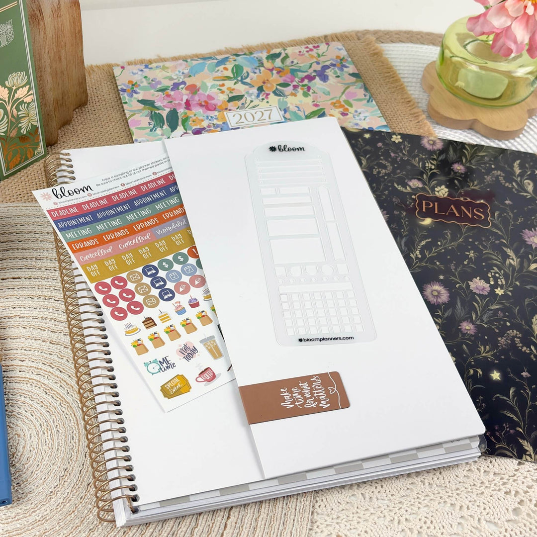 A styled flat-lay of a planner set on a woven mat, featuring a white spiral-bound planner with a clear calendar sticker sheet, colorful weekly label stickers, a floral 2027 planner, and a dark floral 'PLANS' notebook.