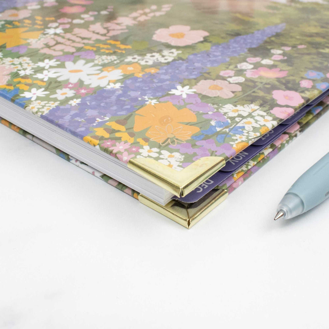 A close-up of a floral-patterned notebook or planner with gold metal corner protectors, shown on a white surface next to a light blue pen.