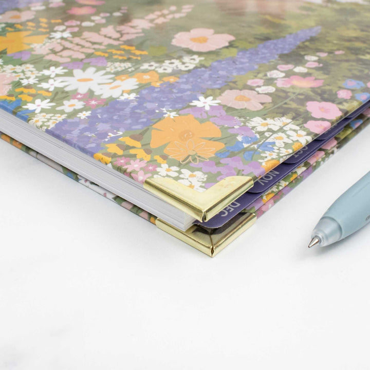 A close-up of a floral-patterned notebook or planner with gold metal corner protectors, shown on a white surface next to a light blue pen.