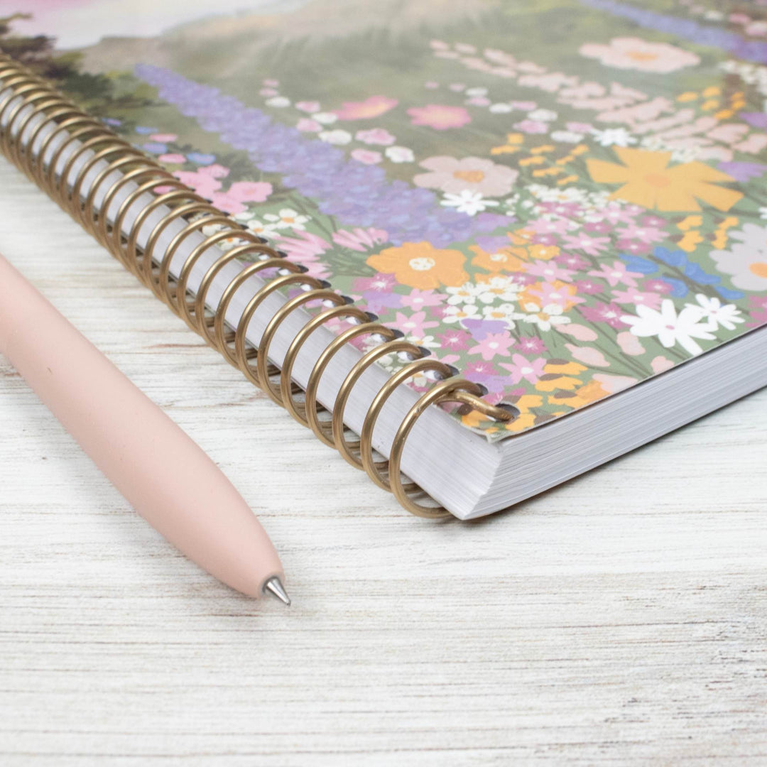 A floral-patterned spiral notebook with a gold-tone wire binding lies on a light wood surface, with a pale pink pen placed diagonally across the foreground.
