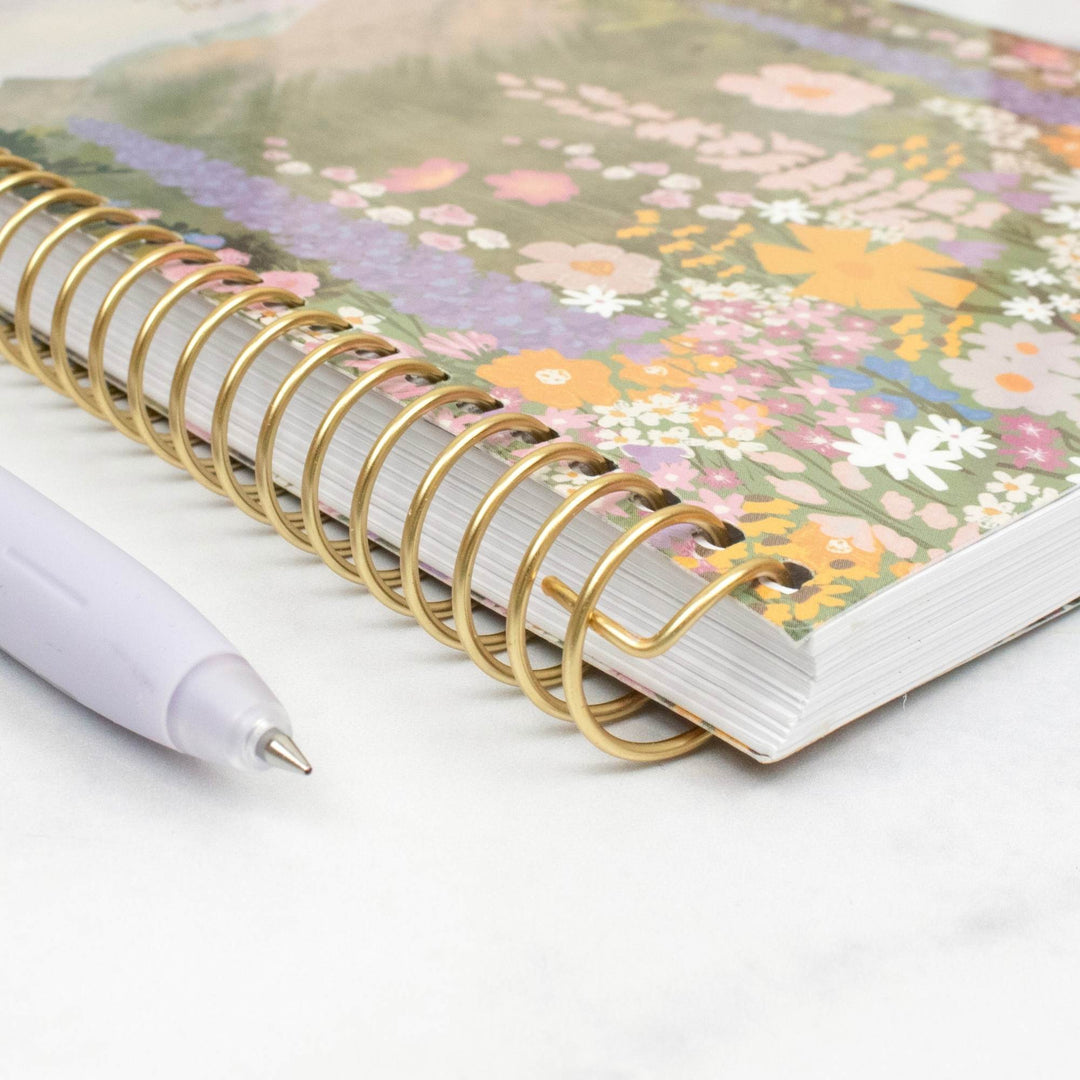 A spiral-bound notebook with a colorful floral cover and gold wire binding, shown on a light marble-like surface next to a white pen.