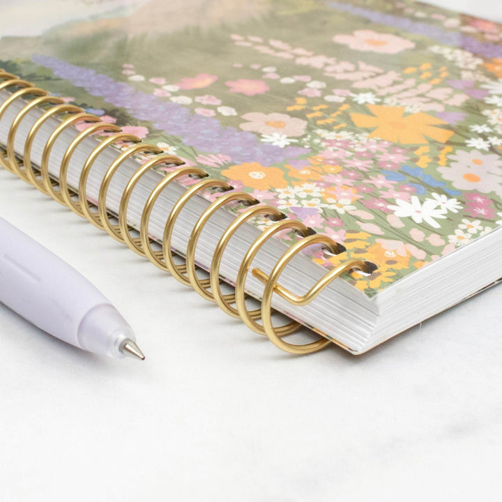 A spiral-bound notebook with a colorful floral cover and gold wire binding, shown on a light marble-like surface next to a white pen.