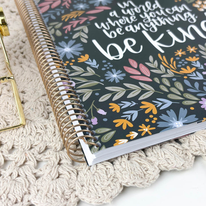 A spiral-bound notebook with a dark floral cover and the phrase 'in a world where you can be kind' in white script, styled on a cream crochet blanket with a gold clip partially visible.