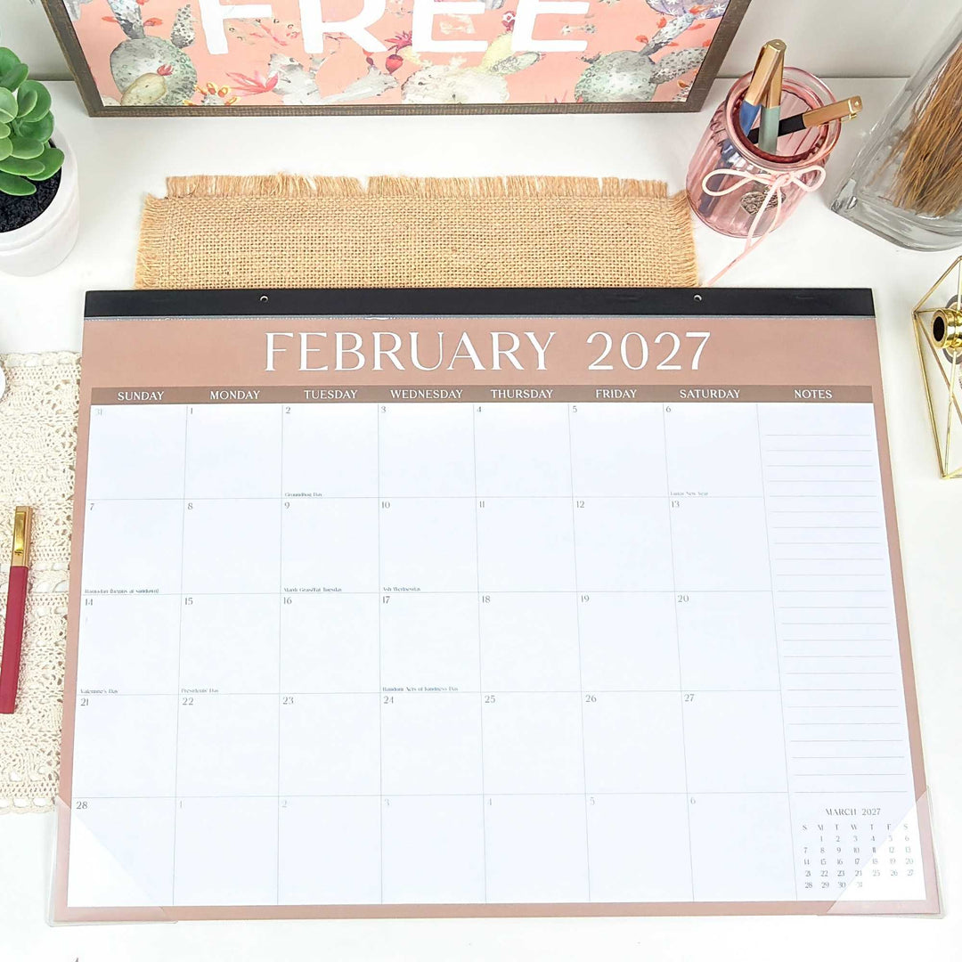 Neutral pink "February 2027" page of a large 16" x 21" desk calendar on a desk. 
