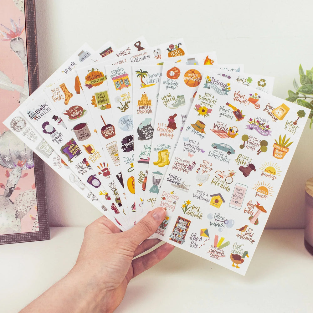A hand holds several bloomdailyplanners Planner Stickers, Seasonal Bucket Lists sheets, each featuring vibrant holiday-themed illustrations and phrases. The colorful sheets are fanned out in front of a wall and a pink potted plant.