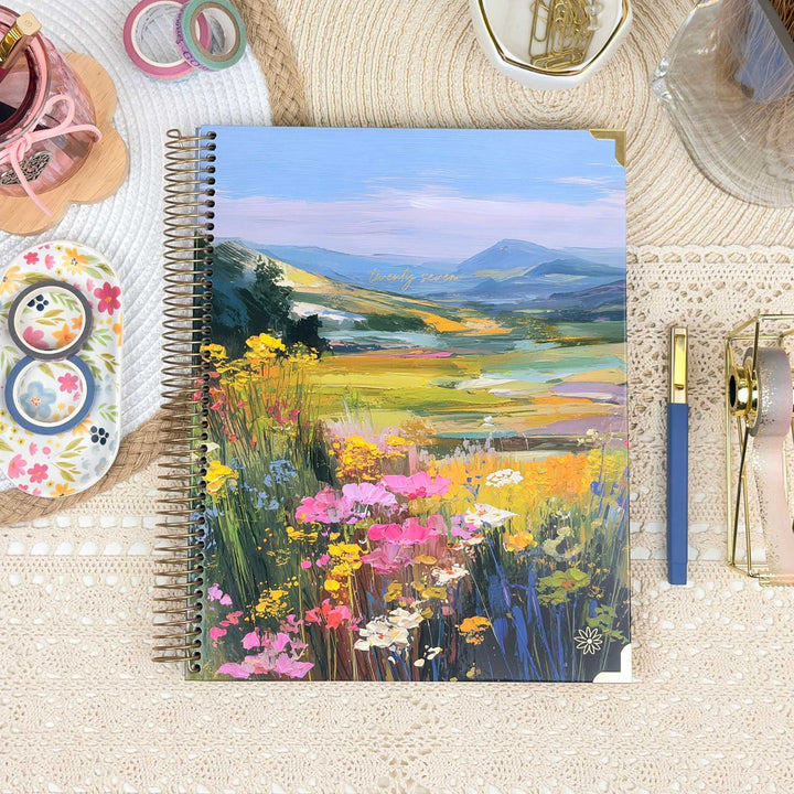 A spiral-bound notebook featuring a colorful landscape painting of a wildflower meadow and distant mountains, styled on a beige lace surface with decorative stationery props.