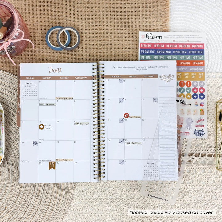 An open spiral planner showing a June calendar page, styled on a woven surface with washi tape, stickers, and a small container. The planner cover reads bloom.