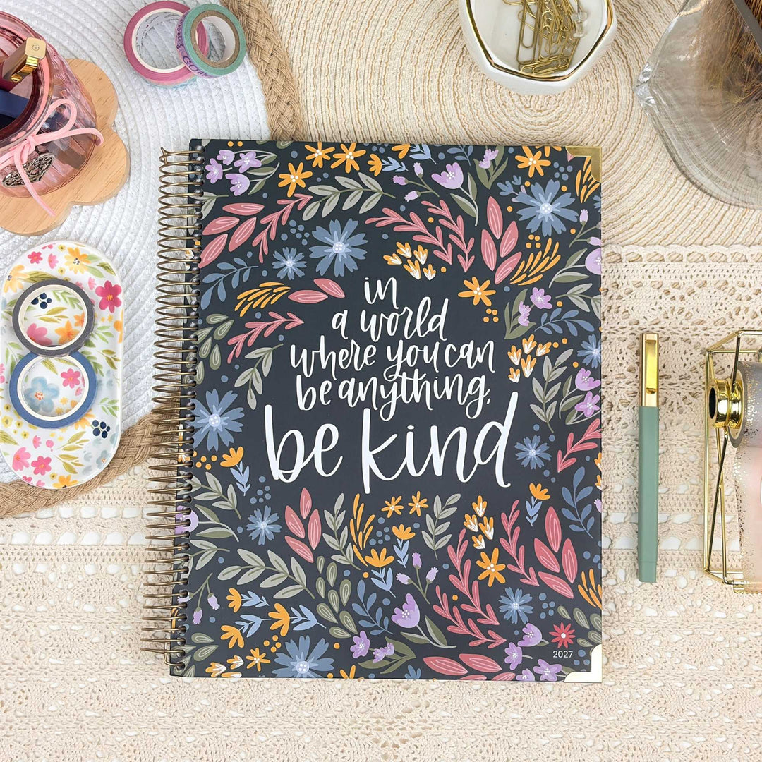 A spiral-bound planner or notebook with a floral cover design and the phrase 'in a world where you can be anything be kind' in white script, photographed from above on a woven mat with stationery props around it.