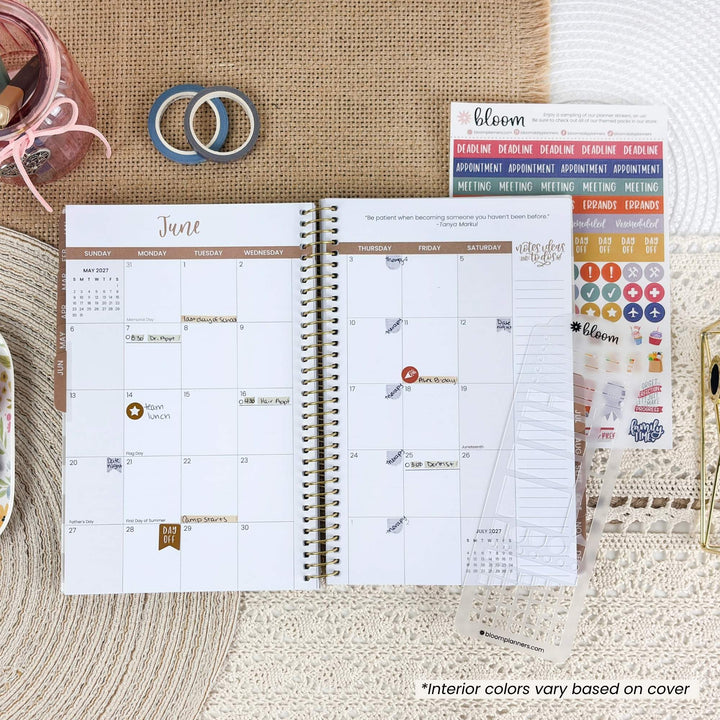 An open spiral planner showing a June calendar page, styled on a woven surface with washi tape, stickers, and a small container. The planner cover reads bloom.