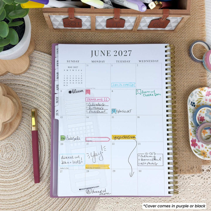 An open spiral planner showing the June 2027 calendar spread, photographed from above on a woven mat with desk accessories around it. The planner pages are white with a purple cover edge and gold spiral binding.