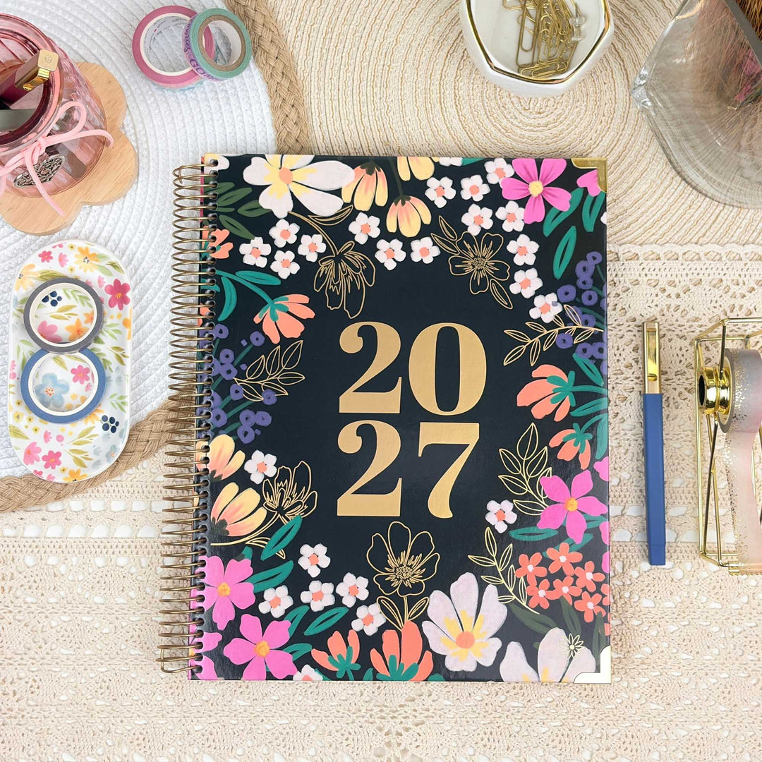 A spiral-bound 2027 planner with a black cover decorated with colorful flowers and gold '2027' text, styled on a beige lace surface with stationery props around it.