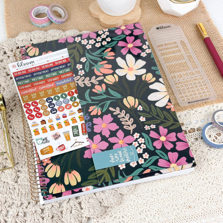 A floral planner cover or notebook with a dark green background and pink, peach, white, and teal flowers, shown with a matching sticker sheet and planner accessories on a textured cream surface.