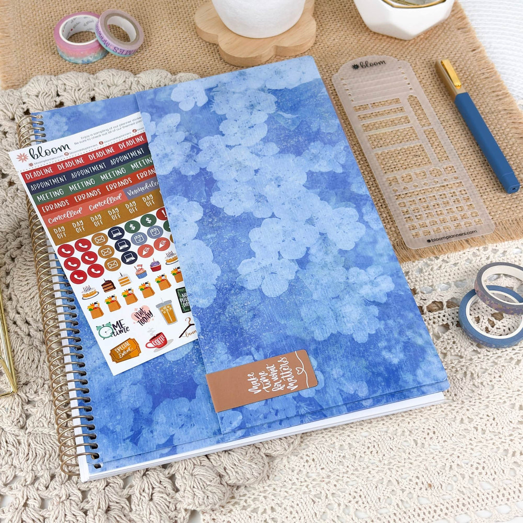 A spiral-bound planner or notebook with a blue watercolor floral cover is shown in a styled flat-lay. A sticker sheet labeled bloom is placed on top, along with a small brown tag reading 'make it beautiful again.