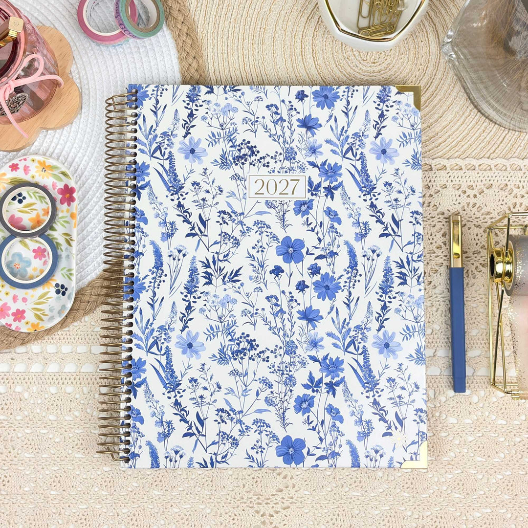A 2027 spiral-bound planner with a white cover decorated with a blue floral pattern, styled on a beige lace surface with stationery accessories.